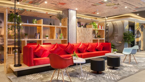 A modern interior with a red sofa, black and red chairs, and a white table. Shelves with plants and books are visible. - Ibis Wuhan Hubu Alley Hotel (Wuhan)