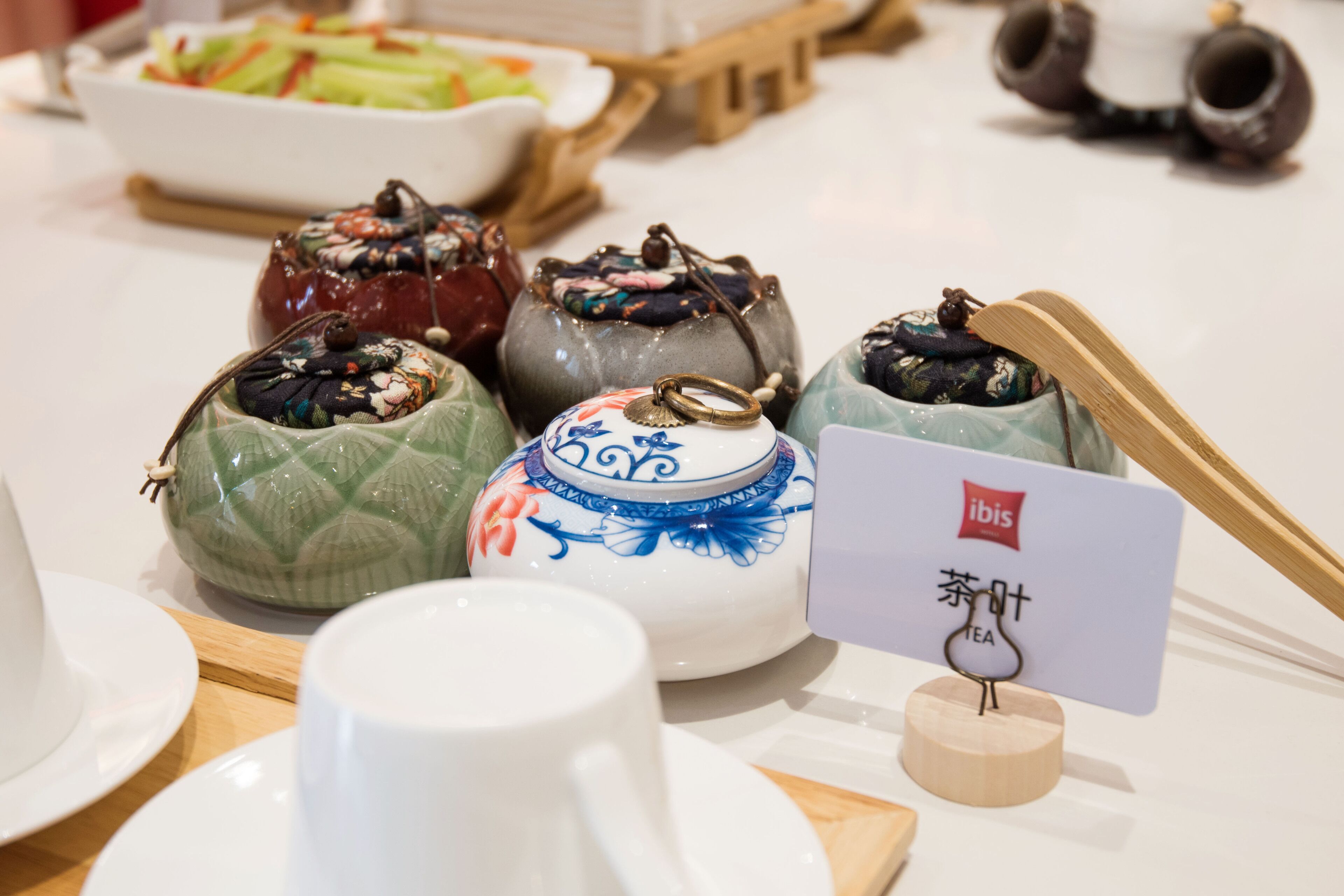 A table with various teapots and a teacup, a sign indicating 'ibis' tea, and a bowl of vegetables in the background.