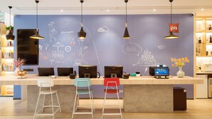 A reception desk with multiple computer monitors, a blue wall with doodles, and three colorful bar stools. - Ibis Xian Xingqing Palace Park Hotel (Xi'an)