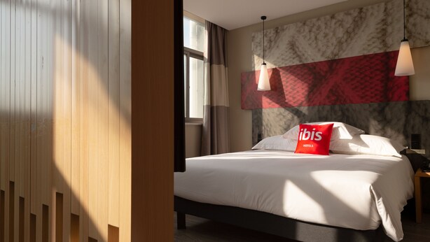 A modern hotel room with a large bed, wooden paneling, a marble accent wall, and a red pillow with the hotel logo.