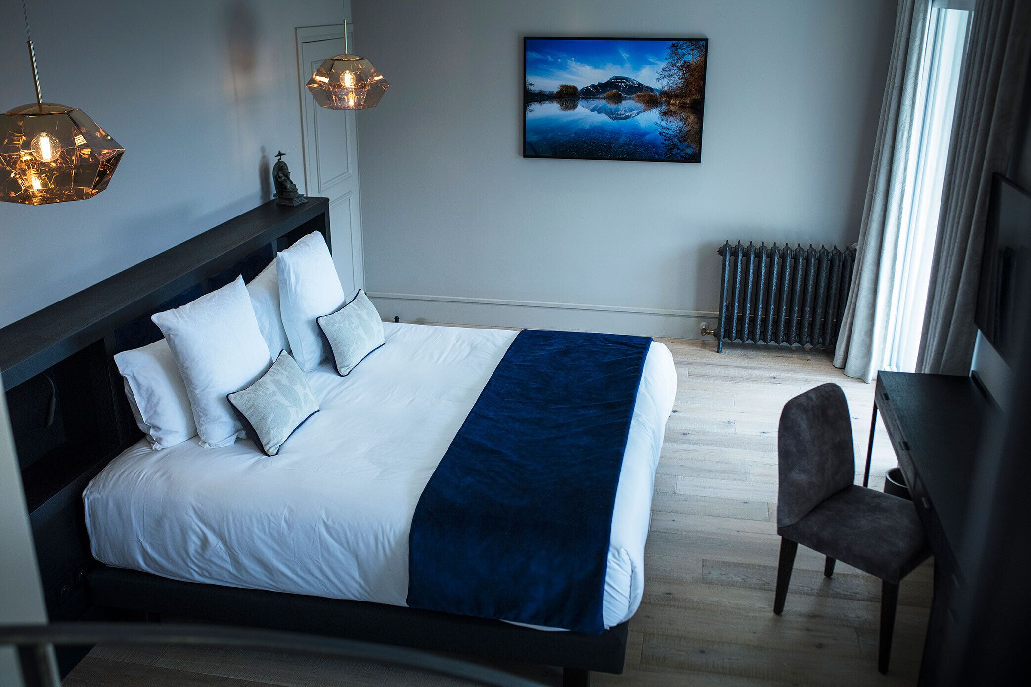 Ein modernes Schlafzimmer mit einem großen Bett, zwei Pendelleuchten, einer Heizung und Blick ins Freie.