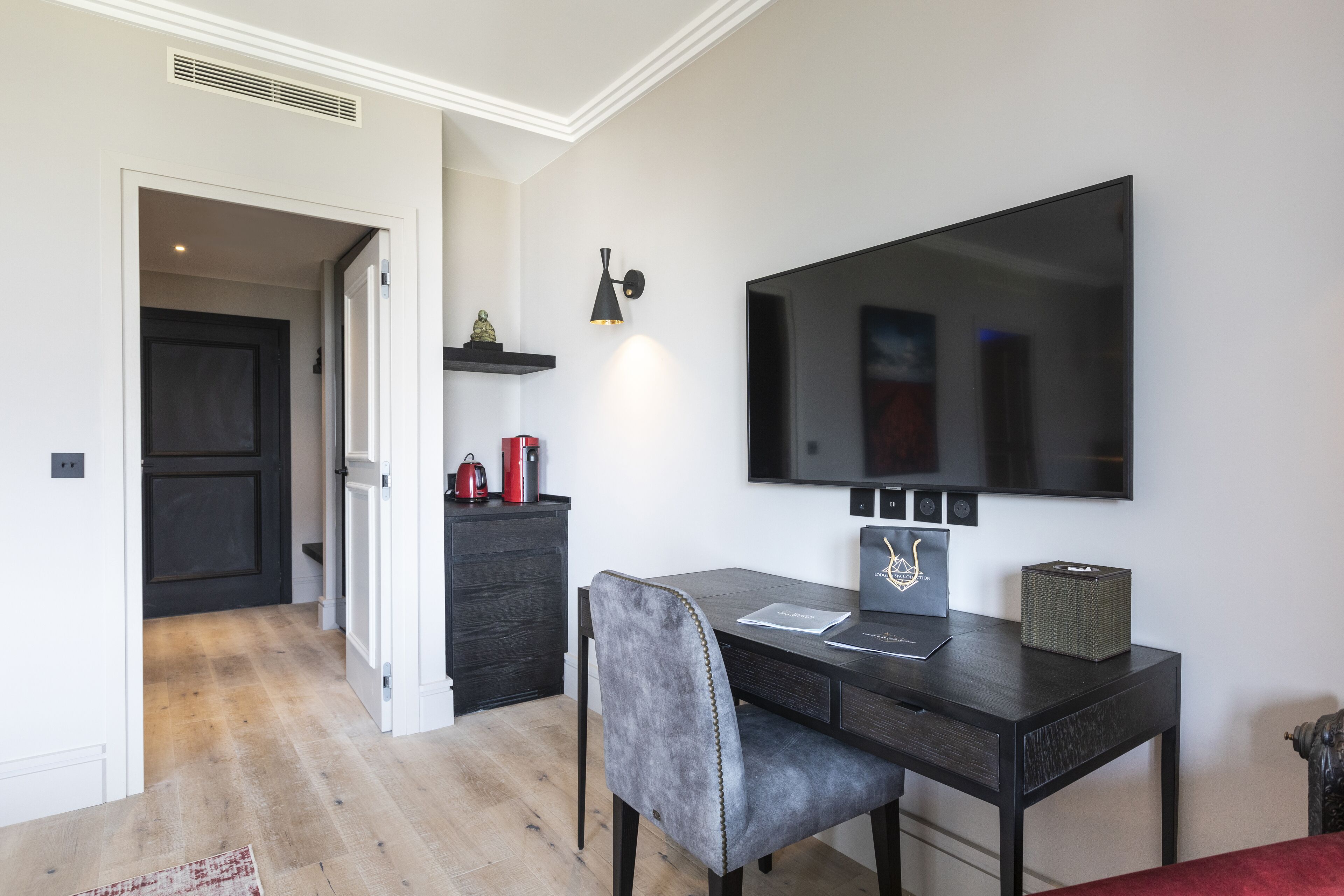 Une chambre moderne avec un bureau en bois foncé, une chaise rembourrée de couleur grise, un téléviseur à écran plat fixé au mur et une petite étagère ornée d’objets décoratifs.