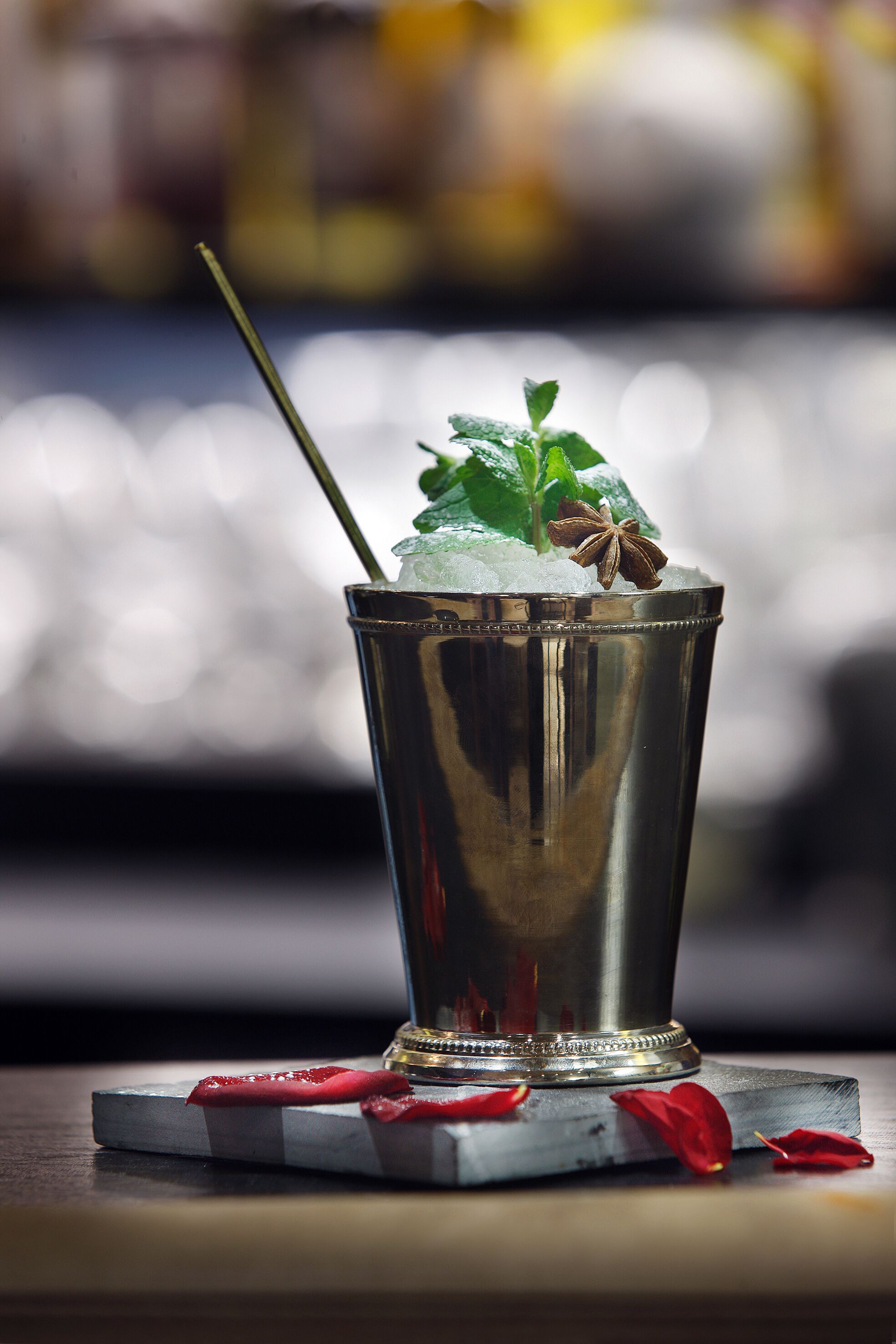 A metallic cocktail glass with a straw, mint leaves, and star anise on ice, placed on a coaster with red petals.