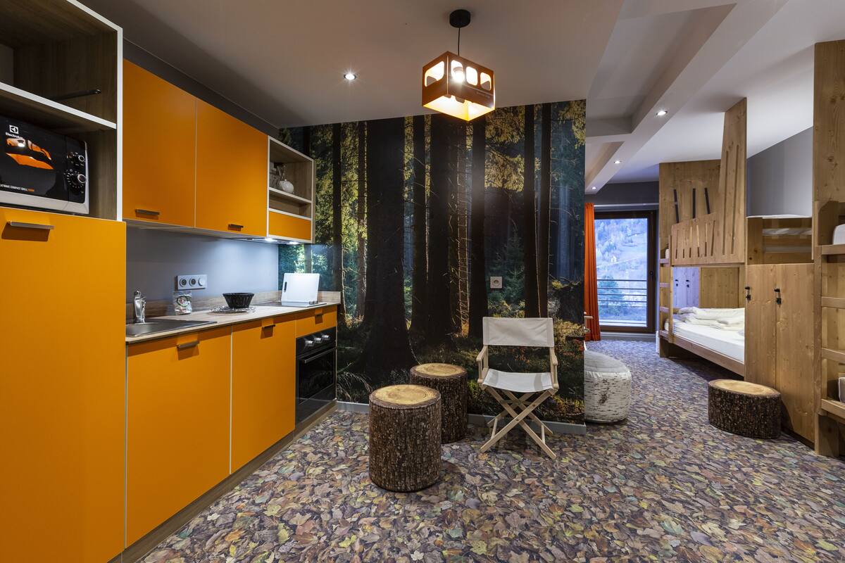 Une cuisine moderne et compacte, avec des meubles de couleur orange, un coin repas en bois et une fresque murale sur le thème de la forêt.