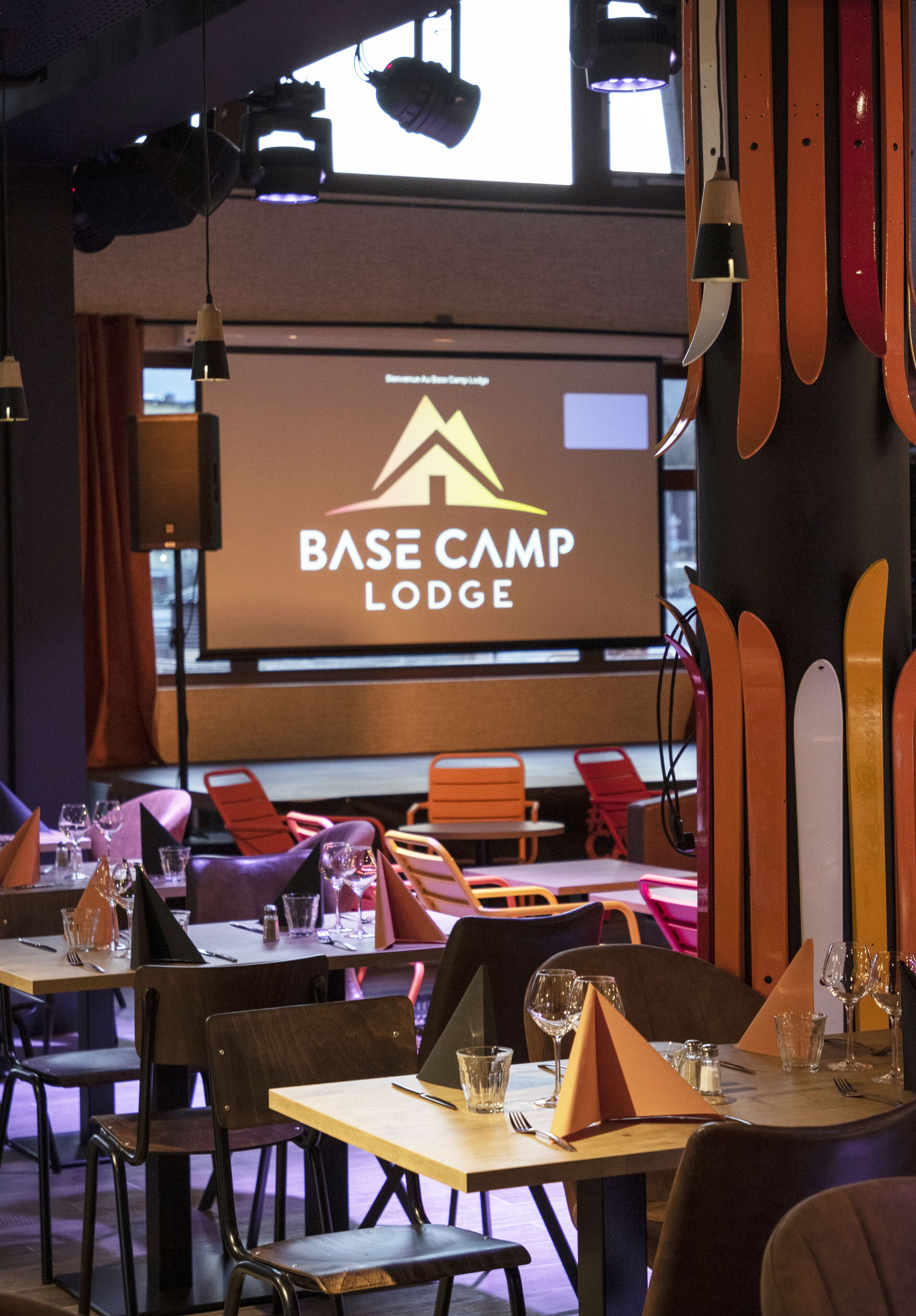 A dining area with tables set for a meal, chairs, and a screen displaying the name "BASE CAMP LODGE".