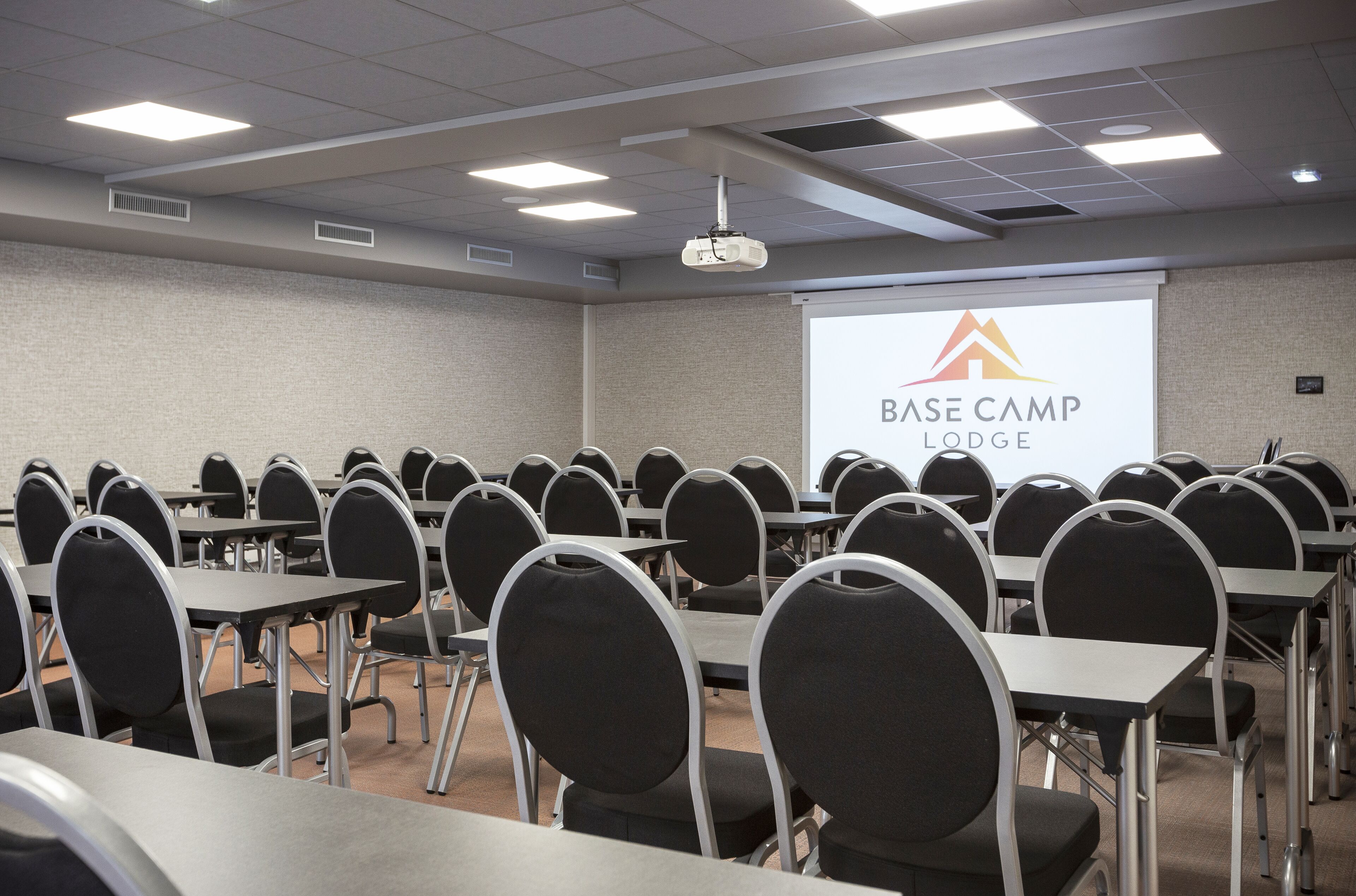 A conference room with rows of tables and chairs, a projector screen displaying the logo of Base Camp Lodge, and a ceiling with recessed lighting.