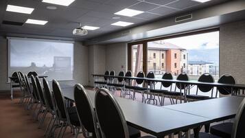 A conference room with a long table and chairs, a projector screen, and a view of buildings through a large window.