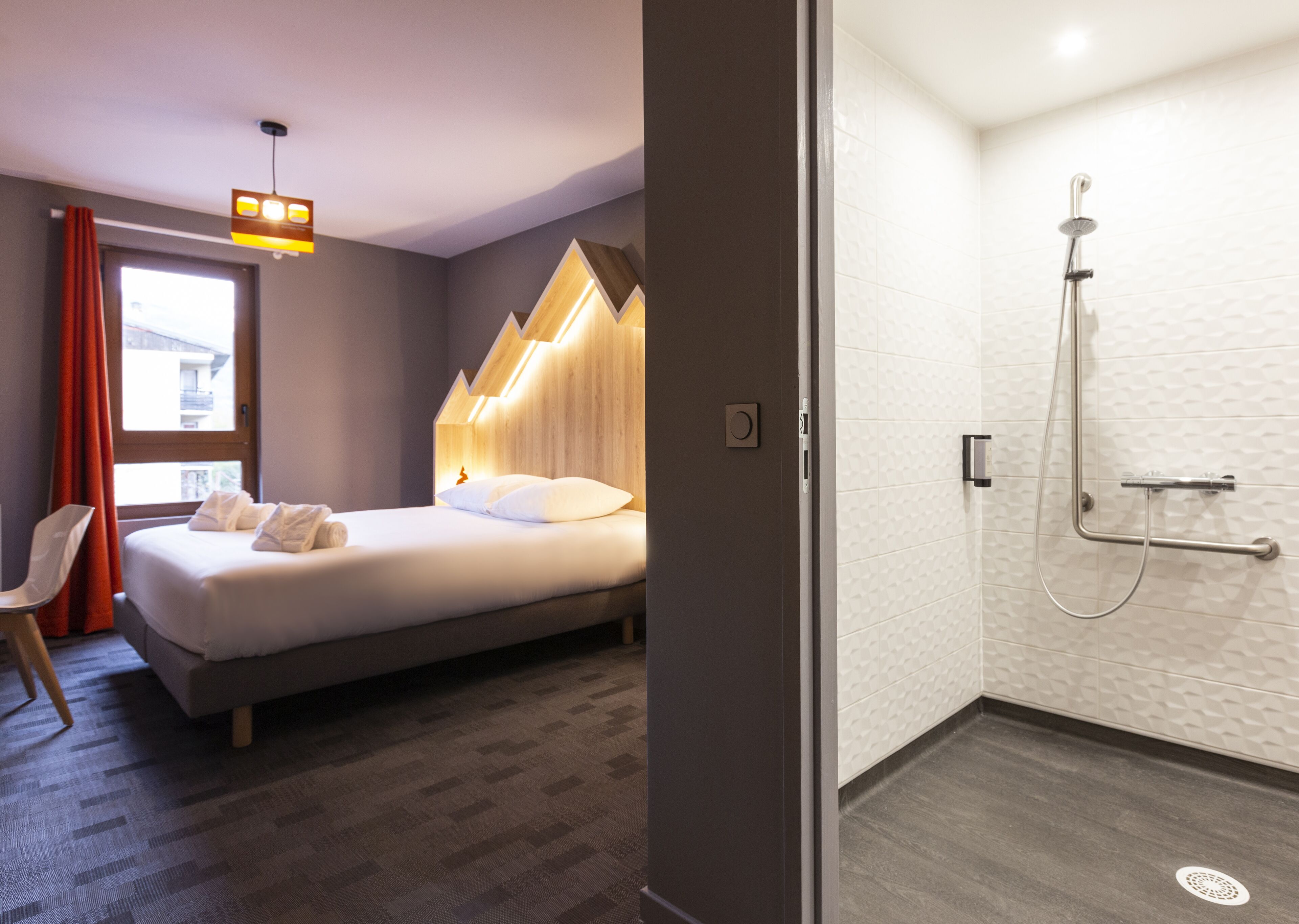 A hotel room with a bed, a chair, a shower area with a handheld showerhead, and a window with curtains.