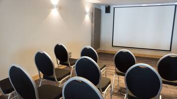 A conference room with rows of chairs facing a projection screen, a whiteboard, and a wall-mounted air conditioning unit.