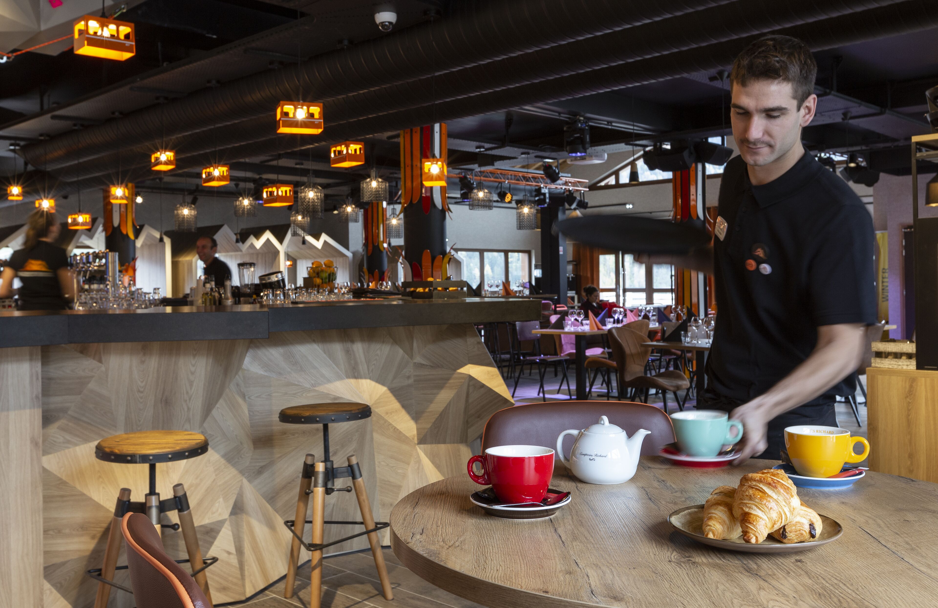 Un café con barra, taburetes de madera y una mesa con un croissant y tazas.