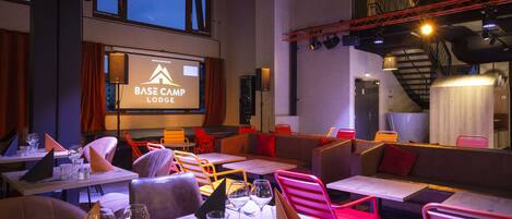 A cozy restaurant with tables, chairs, and a screen displaying "BASE CAMP LODGE".