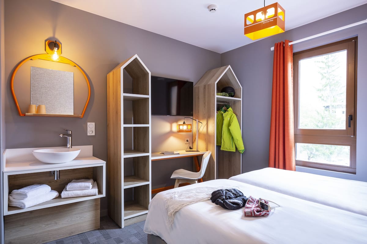 Une chambre d’hôtel moderne avec un lavabo blanc, un grand lit, un bureau avec une chaise et un téléviseur.