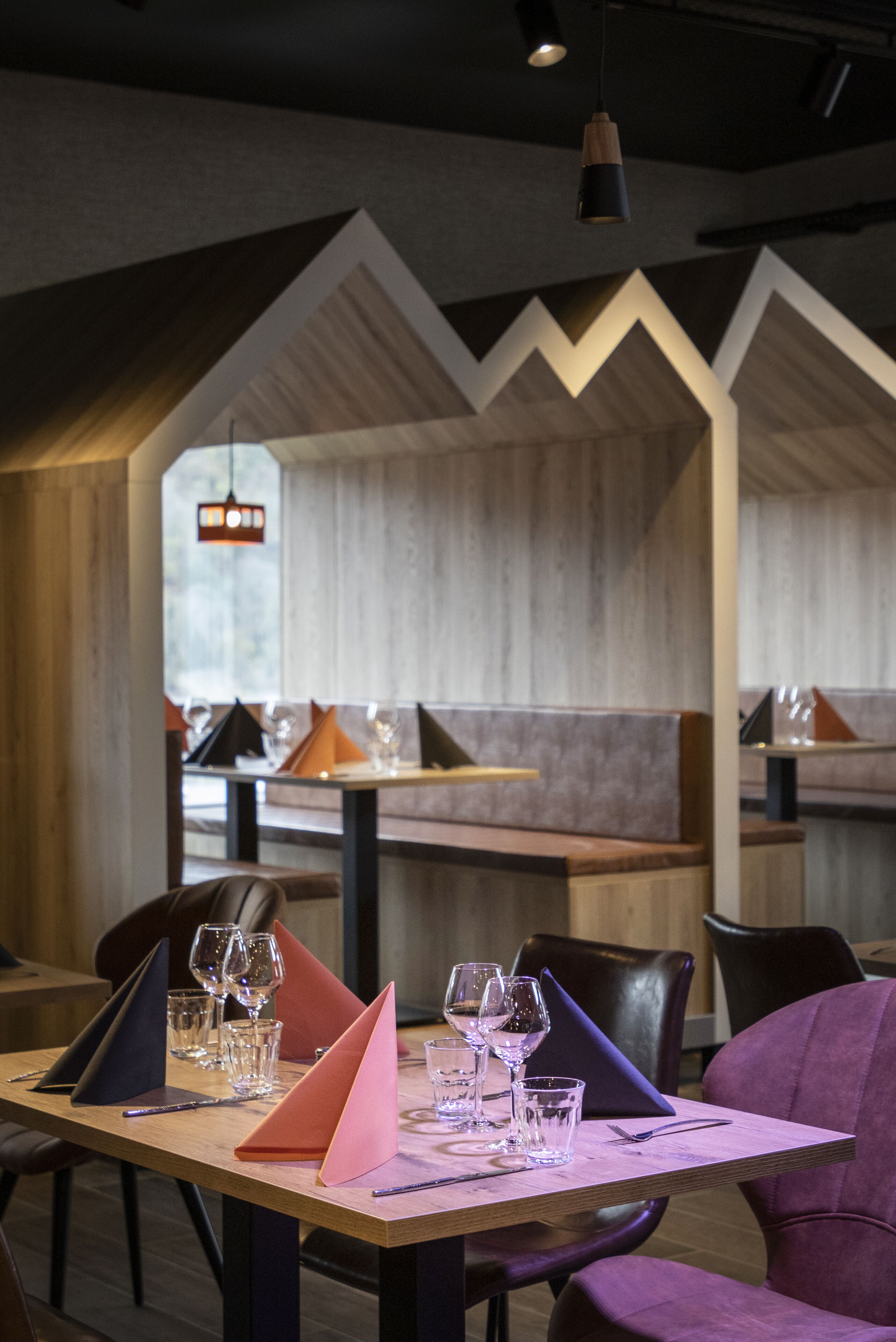 Interior de restaurante con una mesa de madera preparada para comer, una lámpara colgante moderna y una vista del salón comedor a través de un diseño geométrico en la pared.