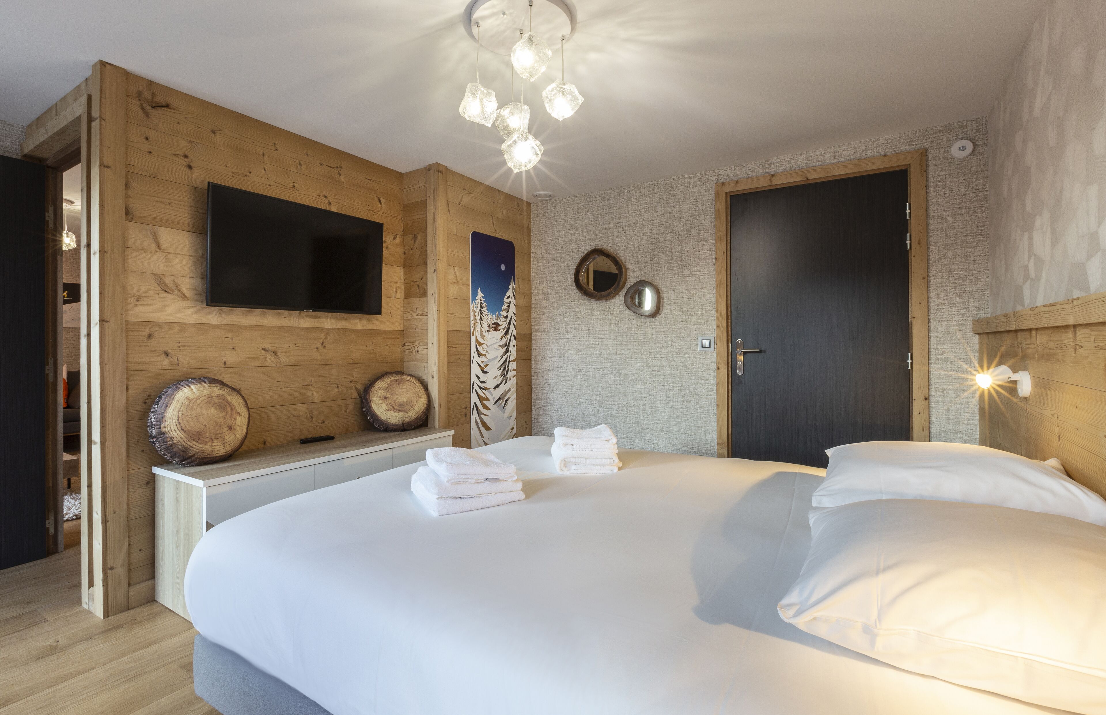 A modern hotel room with a large bed, wooden panel walls, a mounted TV, and a unique chandelier.