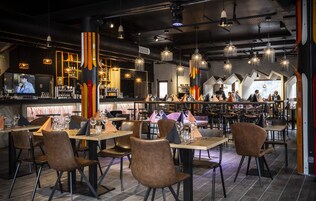 A restaurant with multiple dining tables and chairs, a bar area with a television, and hanging light fixtures.