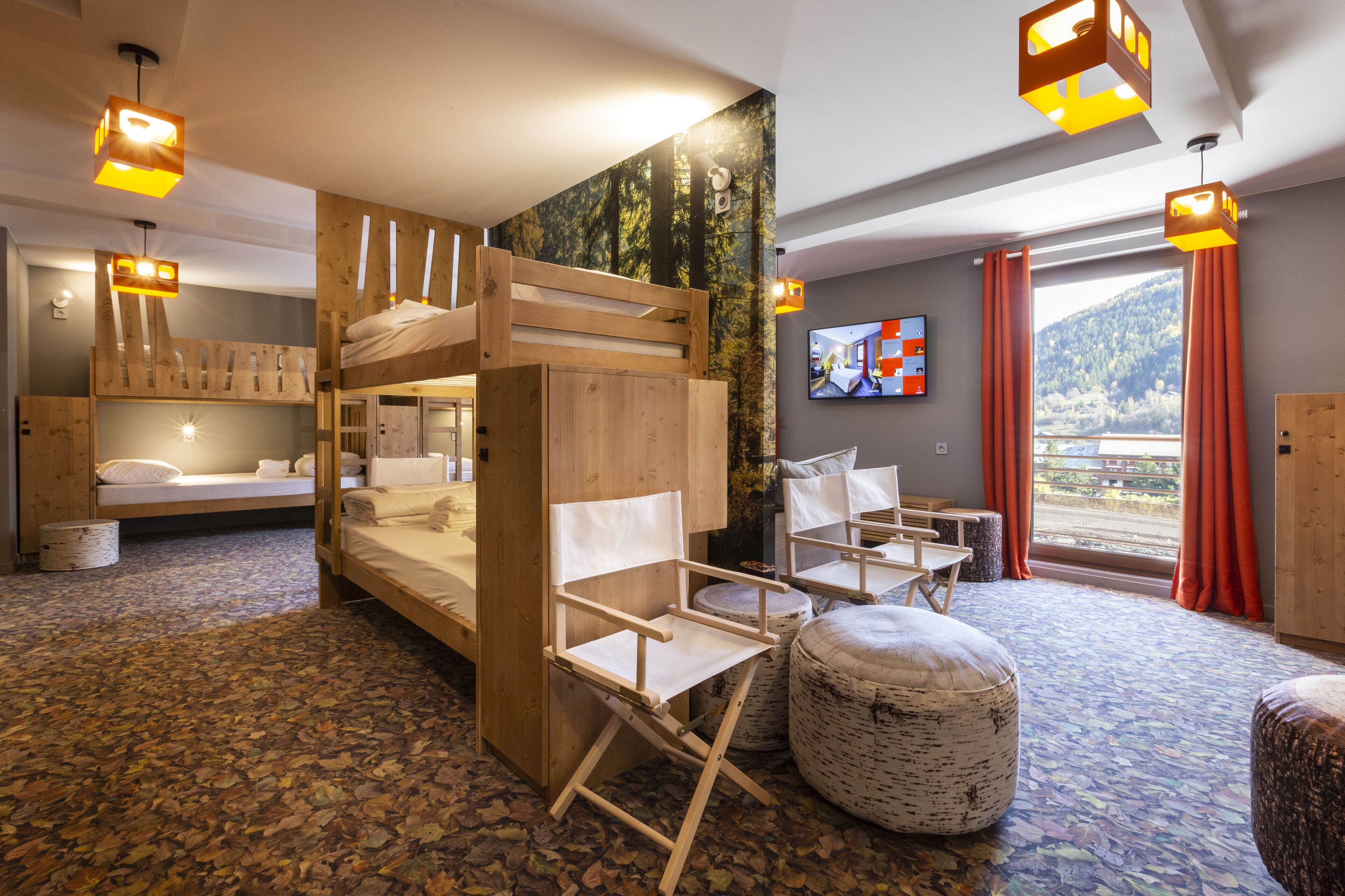 A room with bunk beds, a foldable chair, ottomans, a television, and a view of a mountain.
