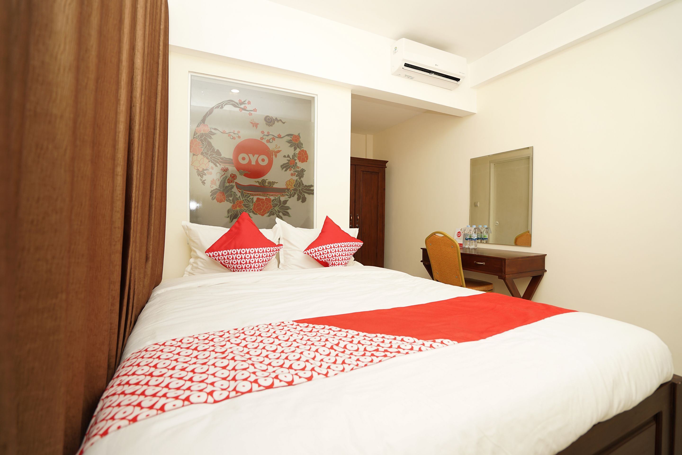 A hotel room with a large bed, a wooden headboard, a desk with a chair, a mirror, and an air conditioning unit.