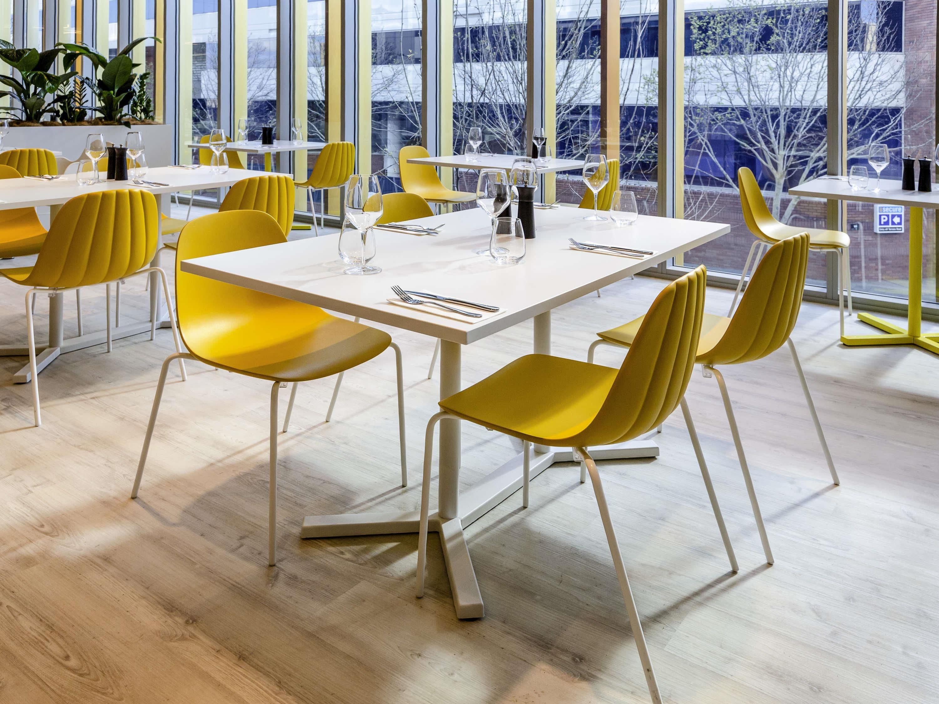 A modern dining area with yellow chairs and white tables, large windows, and a view of the outside.