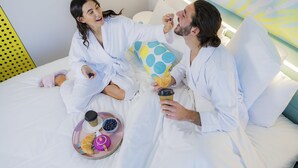 Habitación de hotel con cama, almohadas, una bandeja con comida y bebida, y una pared con un diseño.