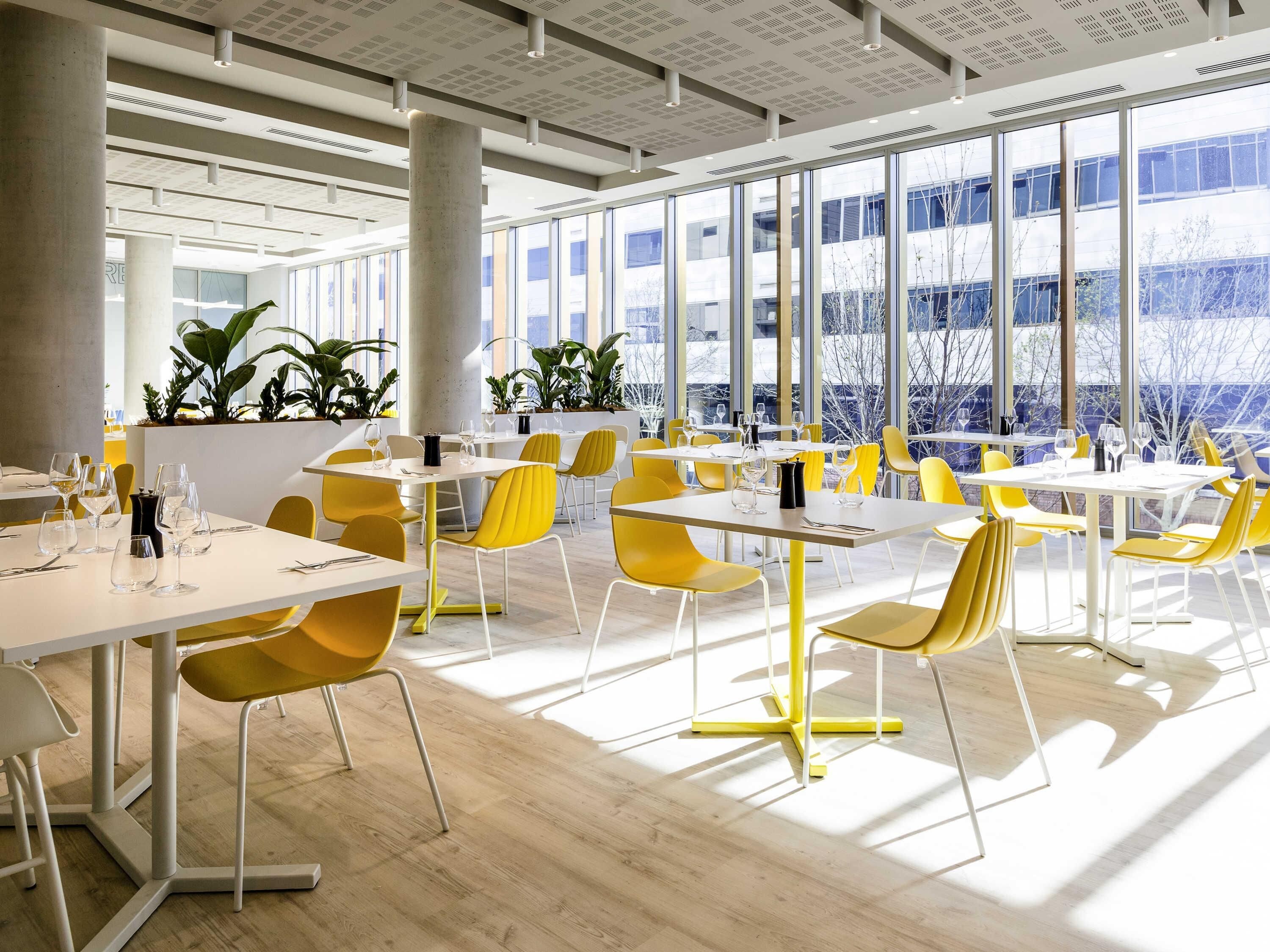 Un comedor moderno con sillas amarillas, mesas blancas y ventanas grandes que ofrecen vistas a edificios y árboles.
