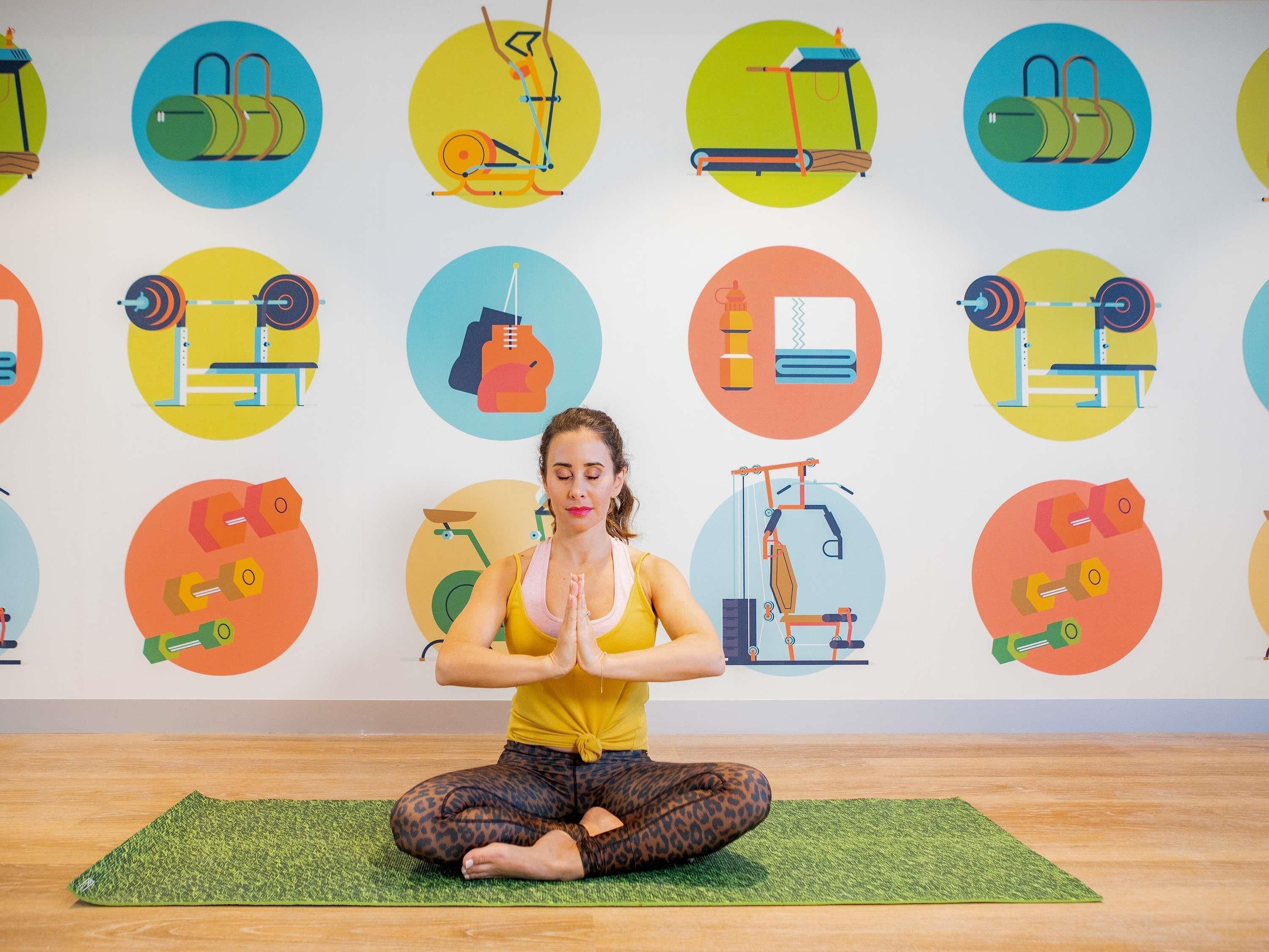 Una persona sentada sobre una colchoneta de yoga en una habitación con una pared decorada con diversas ilustraciones de equipos de gimnasia.