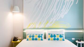 A hotel room with a bed, two pillows with a blue and yellow polka dot pattern, a bedside phone, a small table with a lamp, and a wall with a large abstract painting.