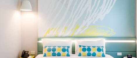 Habitación de hotel con una cama, dos almohadas de estampado azul y amarillo a lunares, un teléfono en la mesita de noche, una pequeña mesa con una lámpara, y una pared con un cuadro abstracto de gran tamaño.