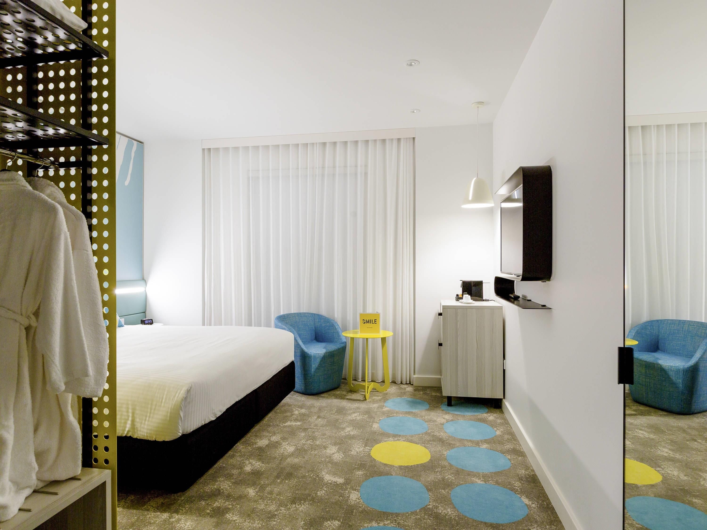 A hotel room with a bed, a mirror, a TV, a small table, and a chair.