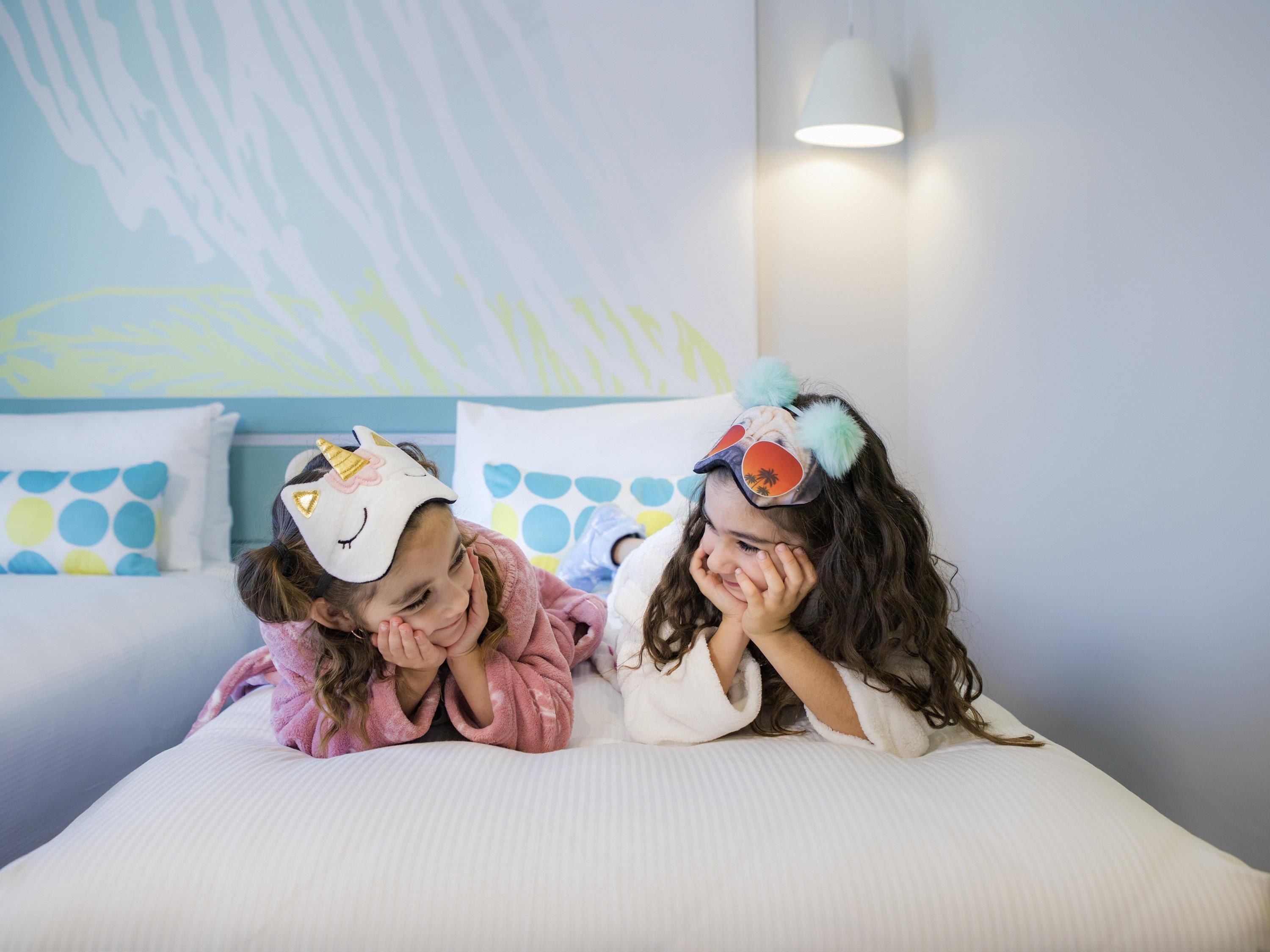 Two children lying on a bed with a colorful wall mural and a white lamp in the background.