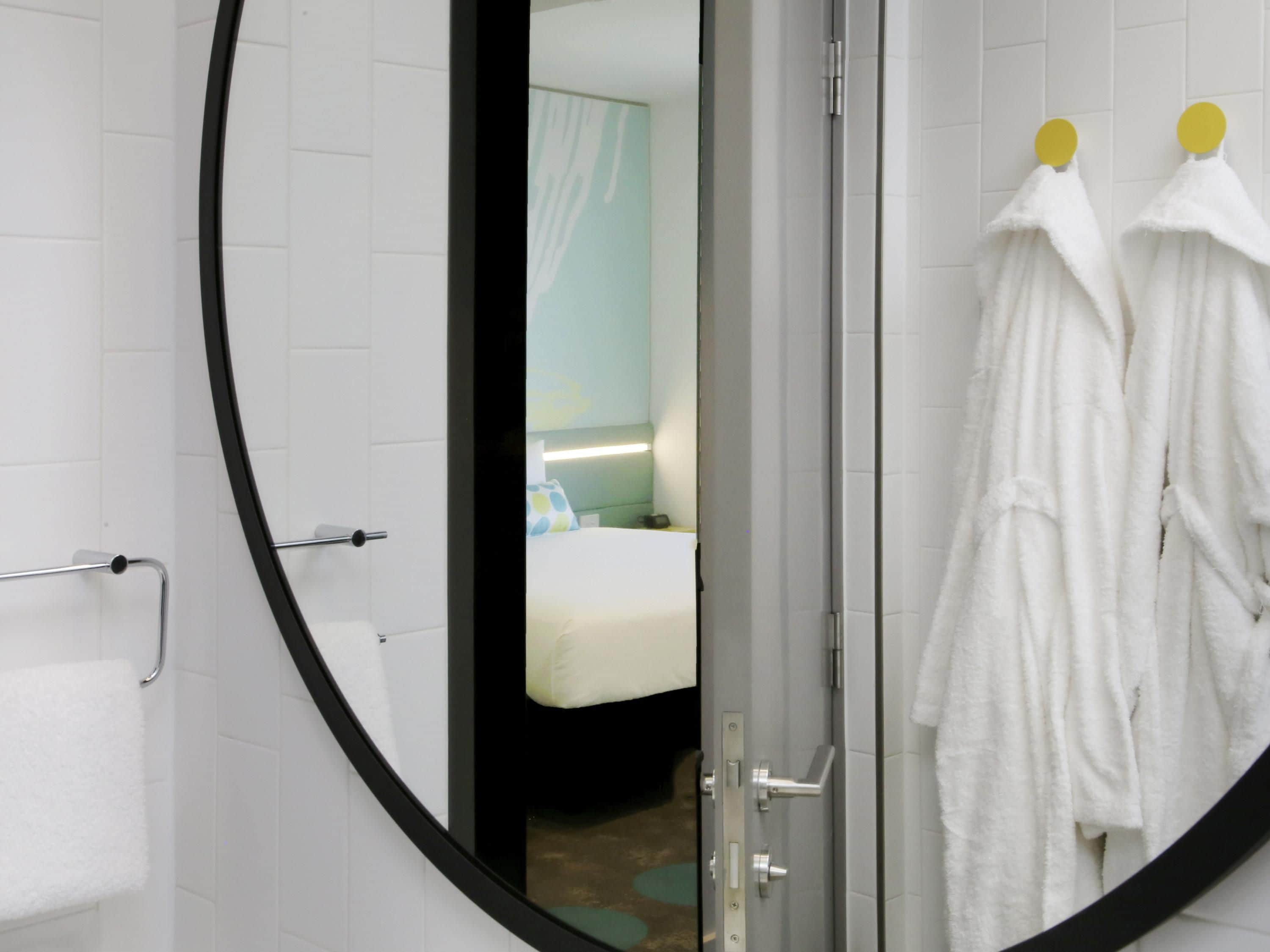 A bathroom with white tiles, a round mirror, and two white bathrobes hanging on hooks.