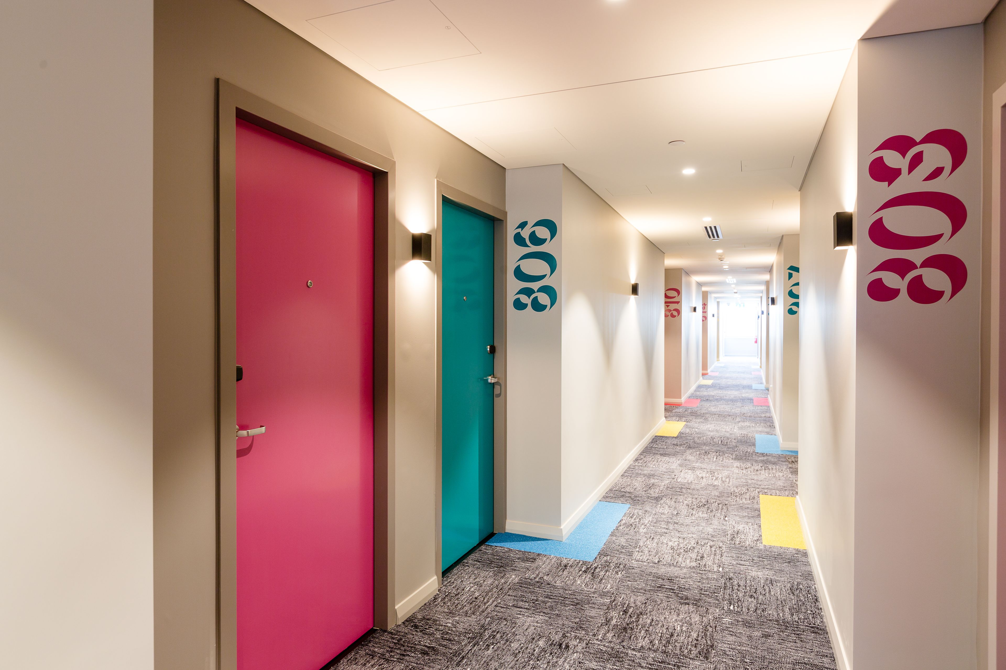 A hotel corridor with colorful doors, carpeted flooring, and wall-mounted light fixtures.