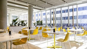 Un comedor moderno con sillas amarillas, mesas blancas y ventanas grandes que ofrecen vistas a edificios y árboles.