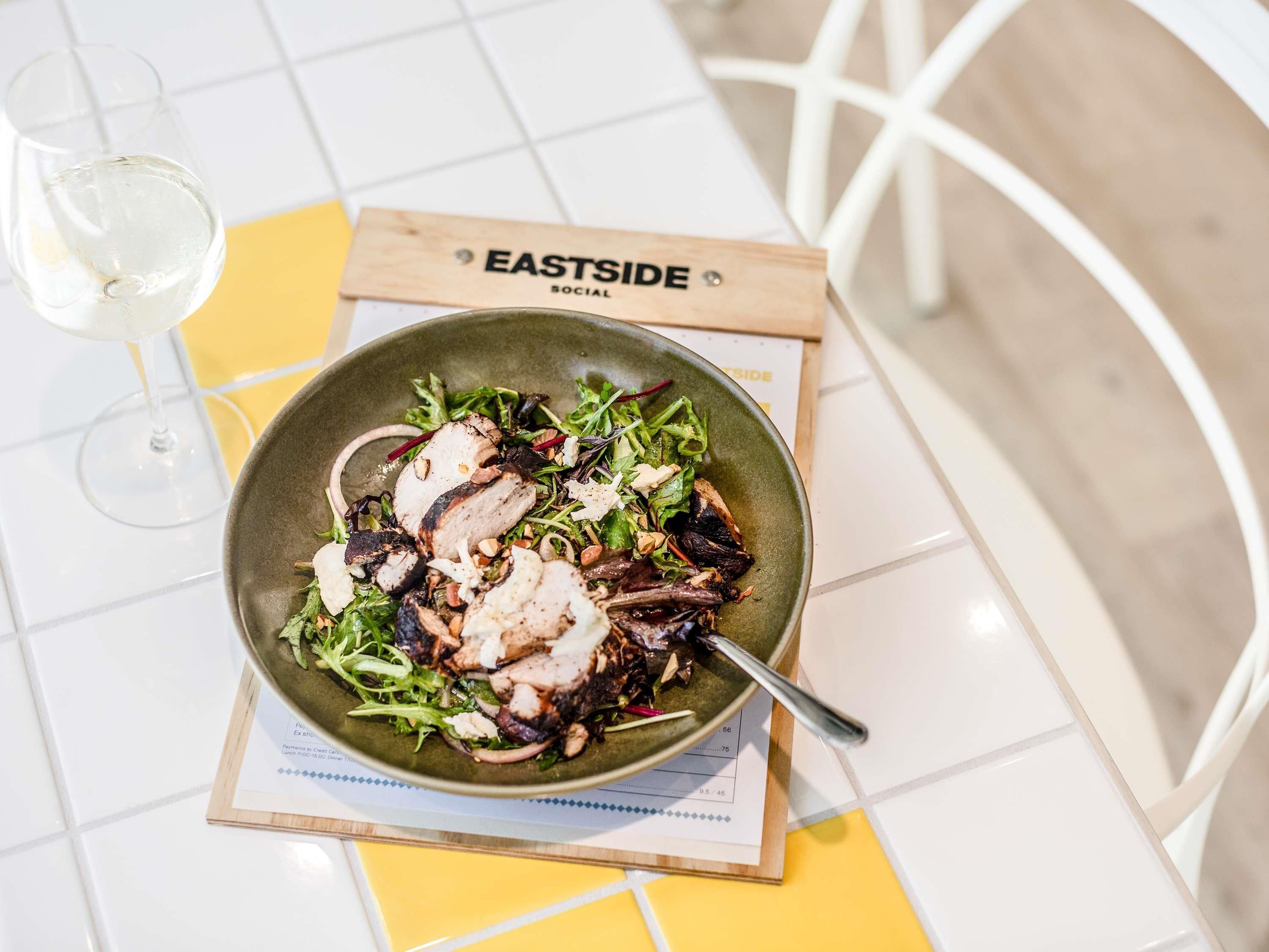 Un tazón de ensalada sobre una mesa con un menú y una copa de vino blanco.