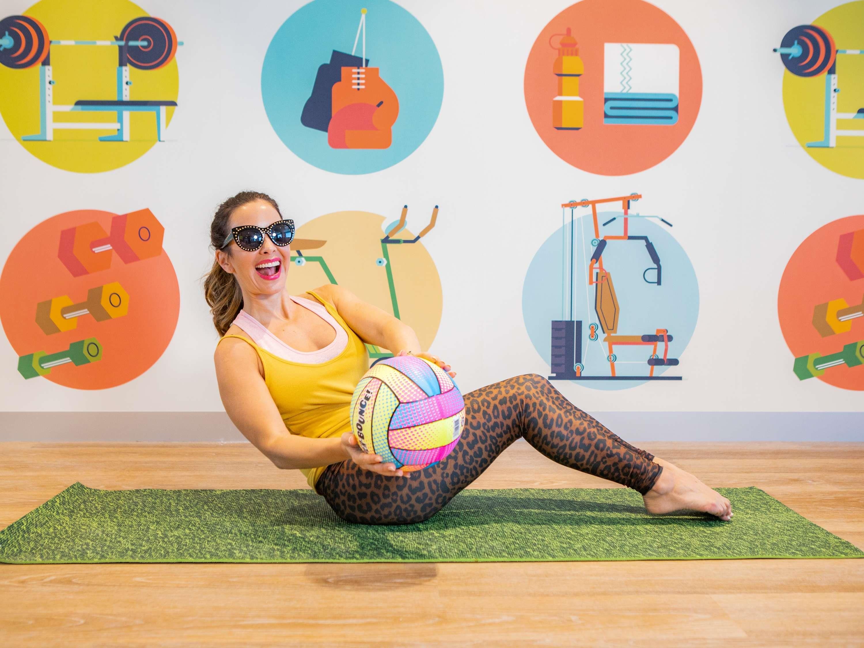 Una persona sentada sobre una esterilla de yoga en una habitación con una pared decorada con iconos relacionados con el ejercicio.