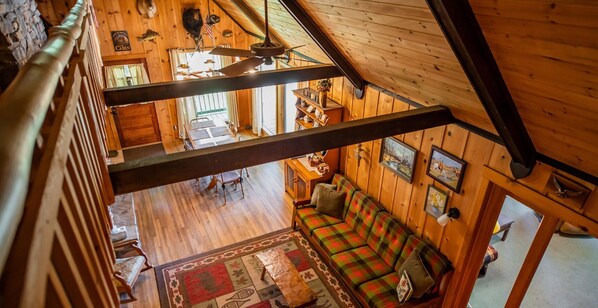 A cabin interior with a wooden floor, a plaid sofa, a ceiling fan, and a dining area with a table and chairs.