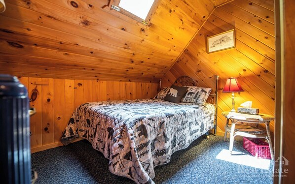 A cozy bedroom with a wooden ceiling, a bed with patterned bedding, a nightstand with a lamp, and a framed picture on the wall.