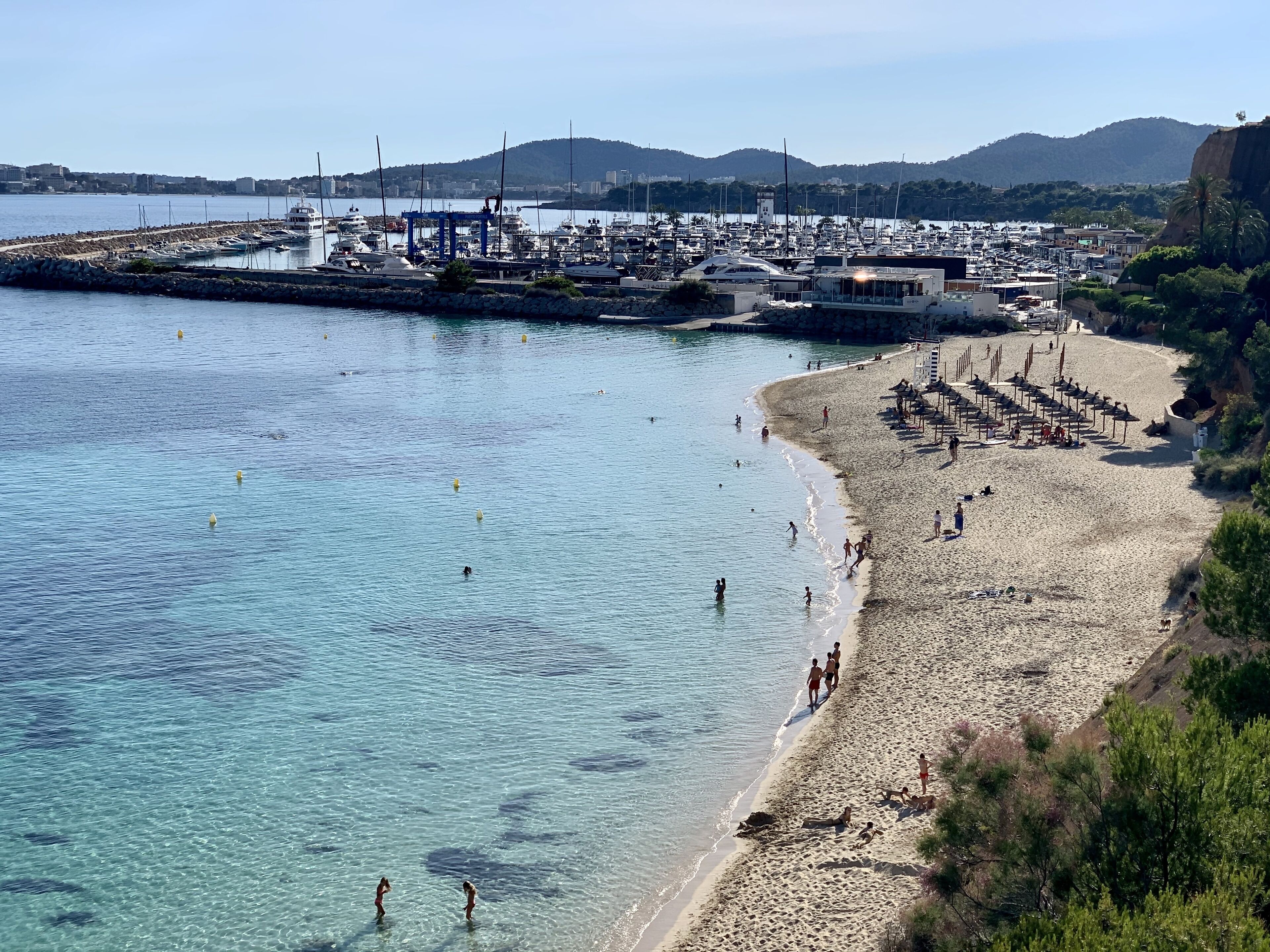 En kystmarina med både, en sandstrand og et bjerglandskab i baggrunden.