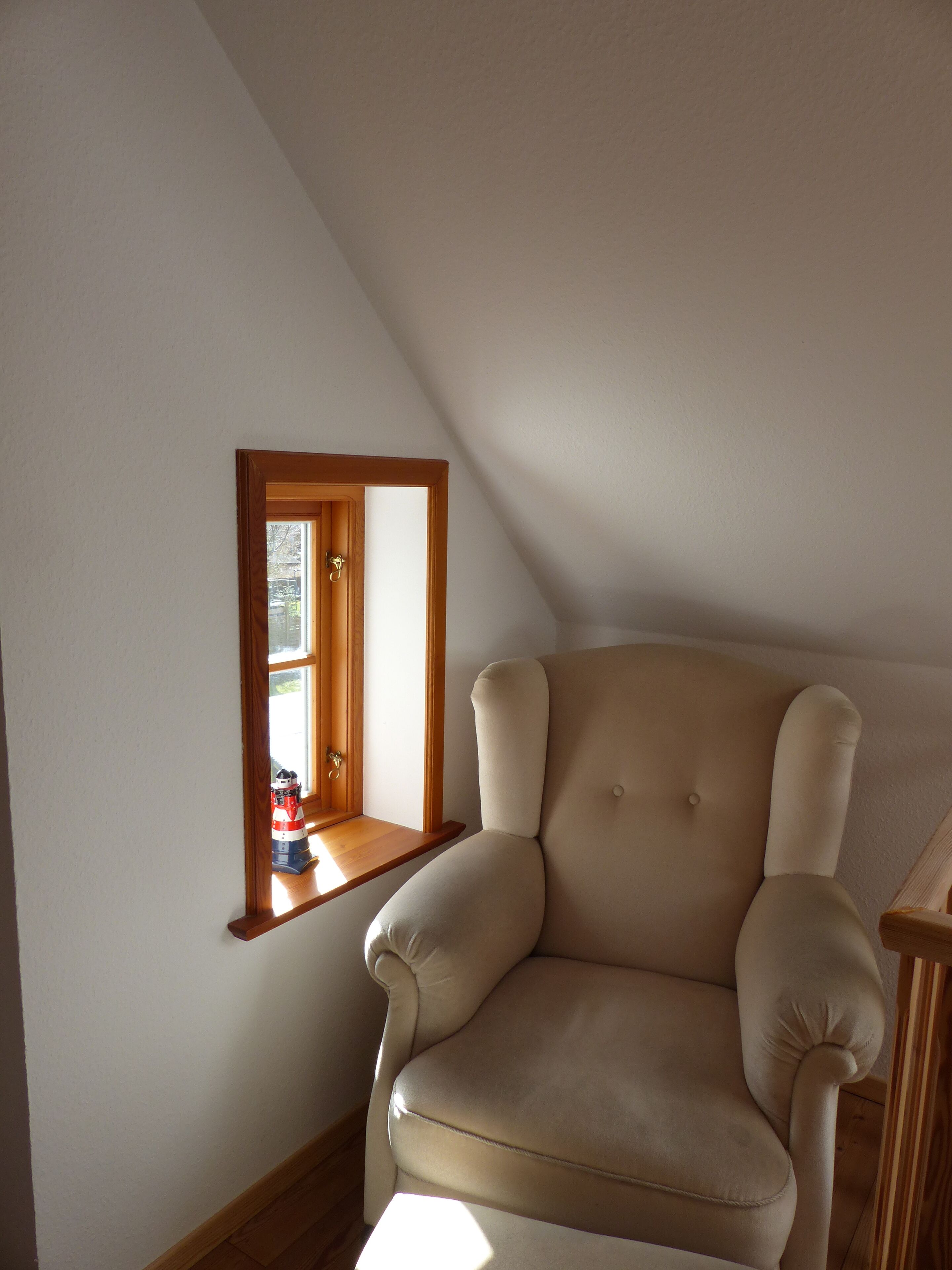 A corner room with a high ceiling, a window with a wooden frame, and a beige armchair.
