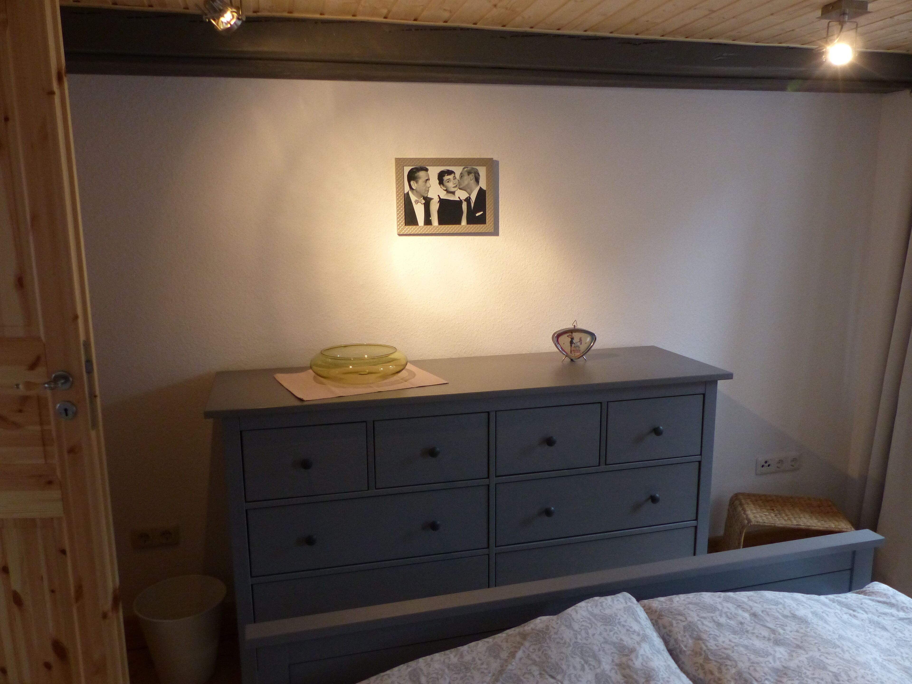 Un dormitorio con cama, cómoda con cajones, taburete pequeño, un cuadro enmarcado en la pared y una lámpara sobre la cómoda.