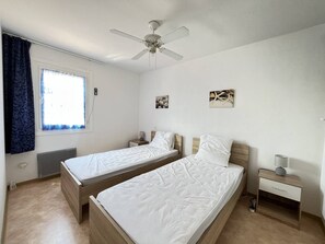 A room with two single beds, a ceiling fan, a window with curtains, and a small bedside table with a lamp.