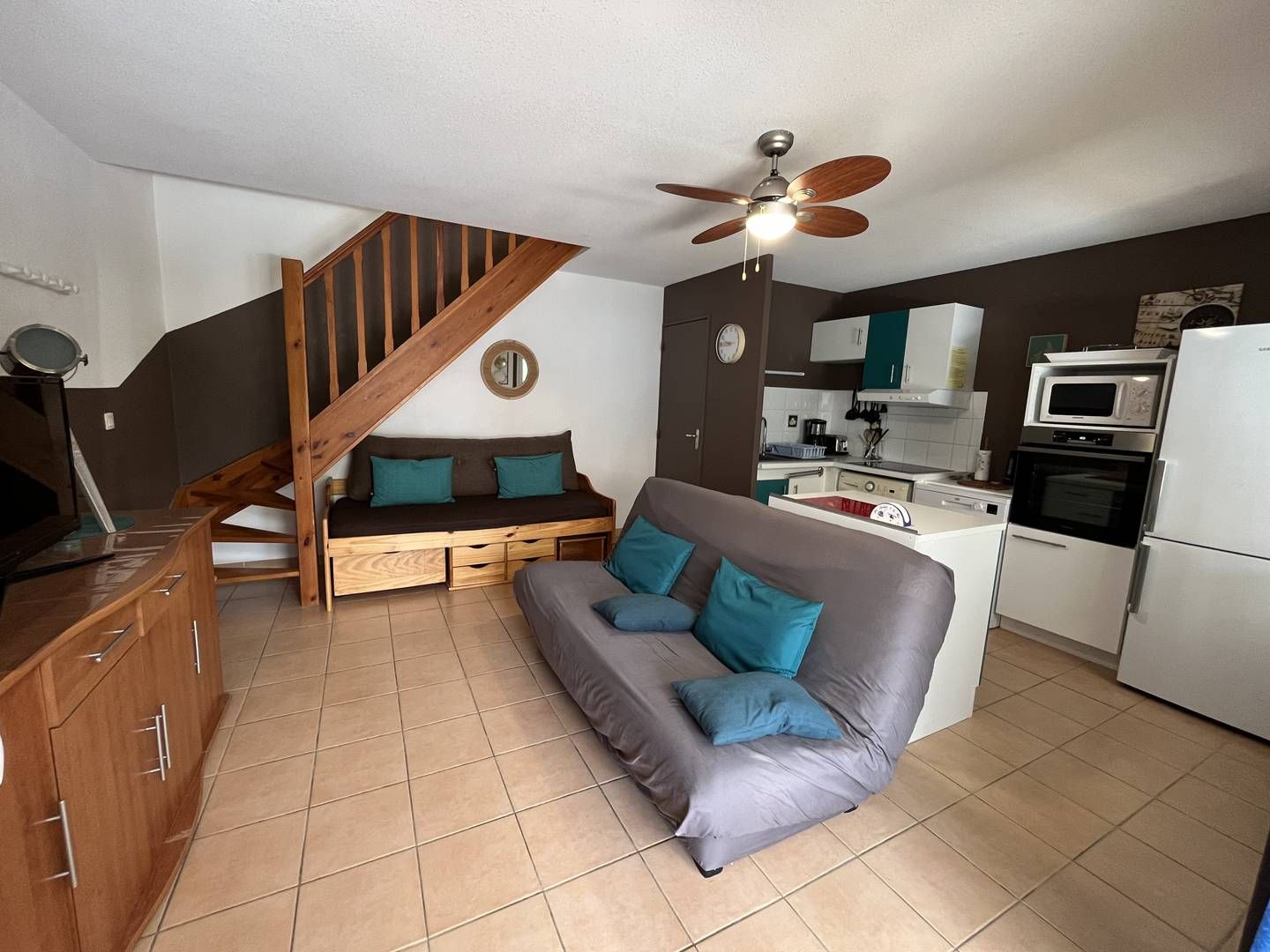 A compact living space with a sofa, a wooden bench, a ceiling fan, a kitchen area with appliances, and a staircase.