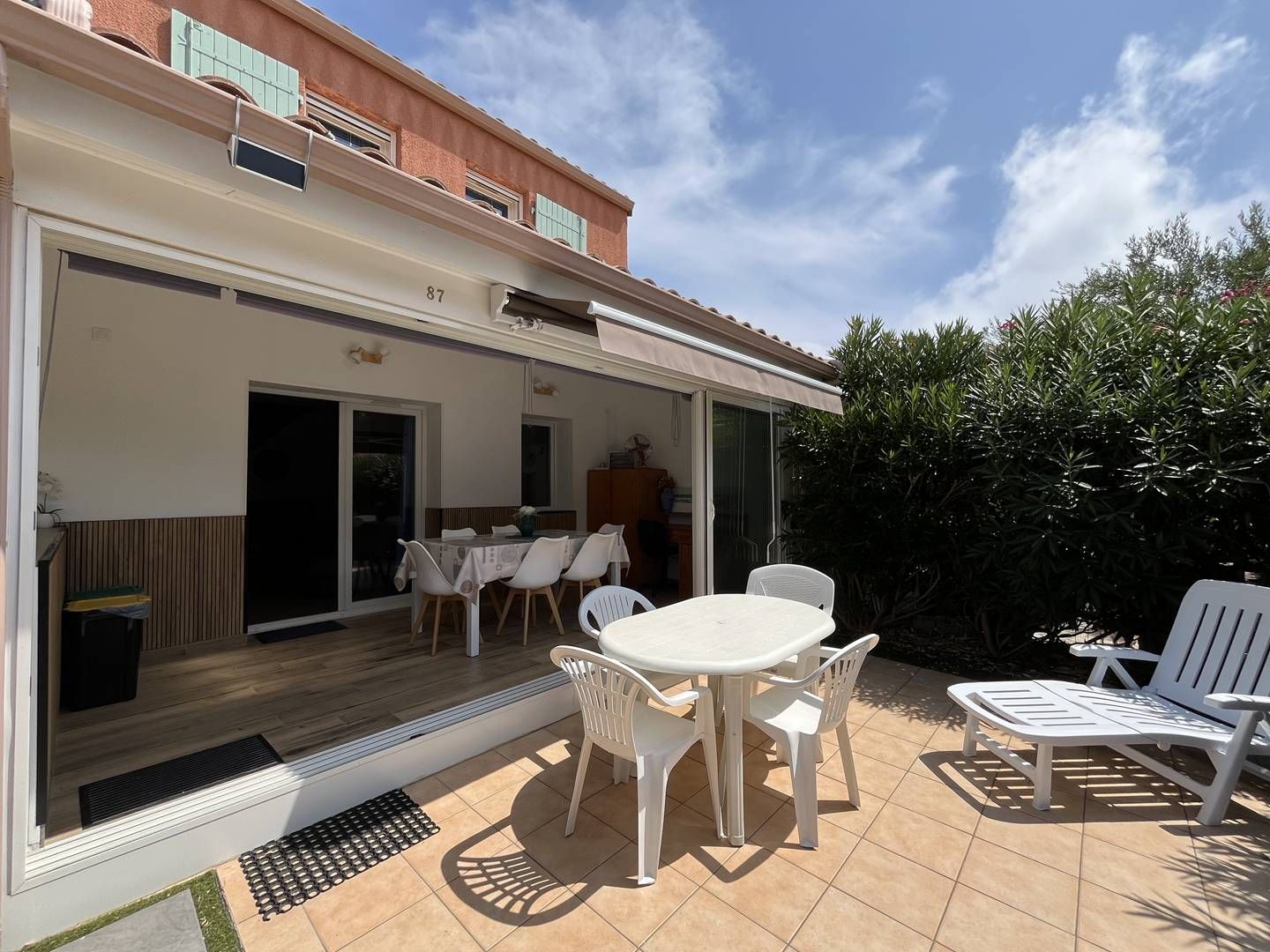 Une terrasse extérieure couverte, dotée d’une table et de chaises, une porte-fenêtre coulissante donnant sur une salle à manger intérieure et un sol carrelé.