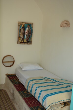 Ein Einzelbett mit einer gestreiften Decke, einem Kissen, einer gewobenen Bank, einem wandmontierten runden Regal und einem Gemälde von zwei Personen.