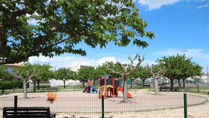 A playground with slides and climbing structures, surrounded by a fence and trees.