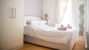 Quarto individual com guarda-roupa, mesa de cabeceira com abajur e janela com cortinas.