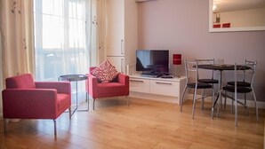 Uma sala de estar moderna com televisão de tela plana, uma área de jantar com cadeiras de metal e um sofá com almofada estampada.