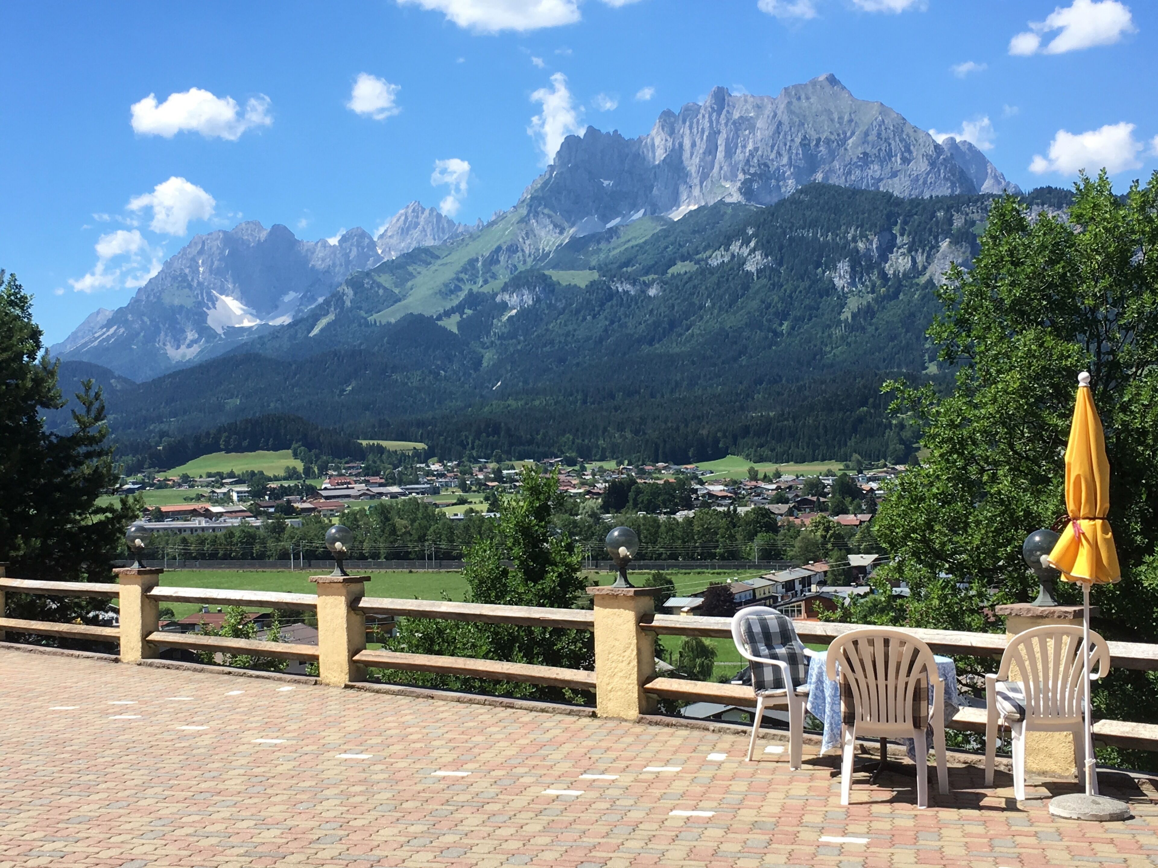 Una terrazza con sedie e un ombrellone giallo, con vista su una città e montagne.
