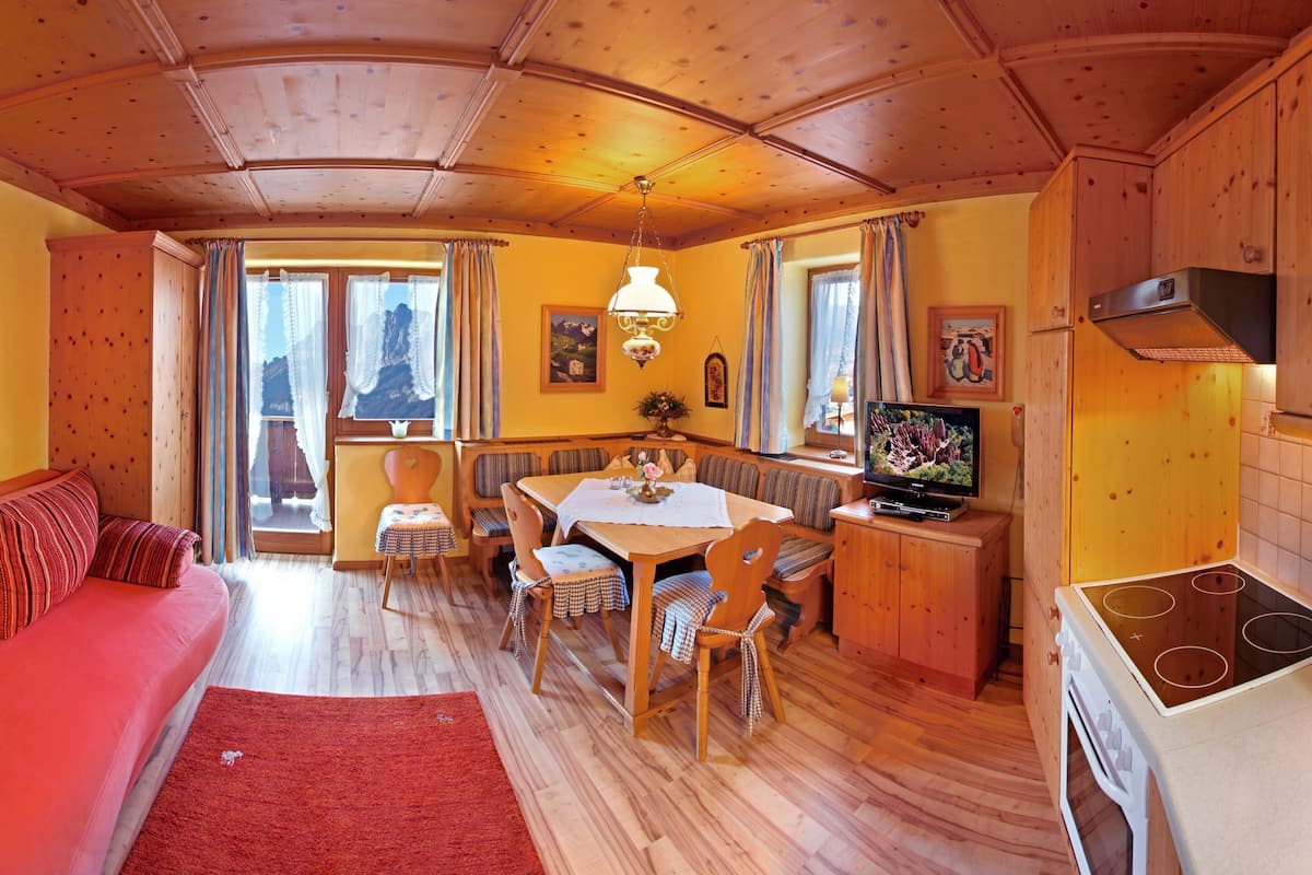A cozy room with a red sofa, wooden dining table, and chairs. There is a television on a wooden cabinet, and a window with curtains.