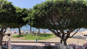 Een restaurant aan het strand met terras, uitzicht op het strand en een grote boom die schaduw biedt.