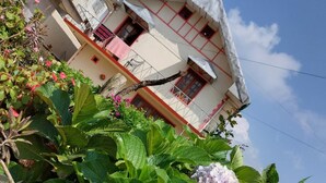 Um edifício com fachada branca e detalhes vermelhos, rodeado por vegetação exuberante e flores de hortênsia.